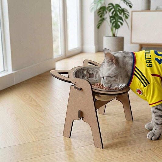 Gato con camiseta comiendo en comedero elevado de madera MDF de Gatitud Store en un espacio interior iluminado.