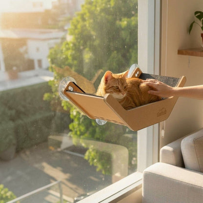 Cama hamaca en V Gatitud Store de madera con tapizado gris instalada en una ventana con ventosas y un gato naranja
